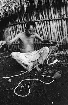 Enfrente de su casa, don Francisco Puc Poot hace una cuerda de fibra de henequén usando su pie como una tercera mano. La cuerda es para sus sandalias que, sin terminar, están junto a su pie izquierdo. Chichimilá, 1971.