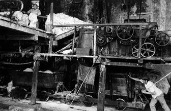 La fibra de henequén sale de la máquina raspadora hacia la plataforma de arriba mientras un trabajador empuja un truc vacío que remplaza uno lleno abajo del conducto que echa los desperdicios de la planta. Ruinas de Aké, 1976.